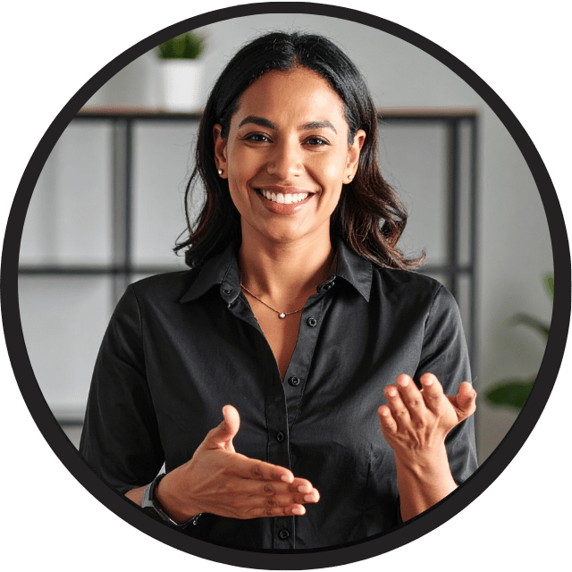 online-asl-interpreter-jobs A person with shoulder-length dark hair is wearing a black button-up shirt and a necklace, standing indoors in front of a shelf with a plant and other items. The individual is gesturing with both hands, suggesting they may be communicating or explaining something, possibly using sign language. Jobs for ASL Interpreters